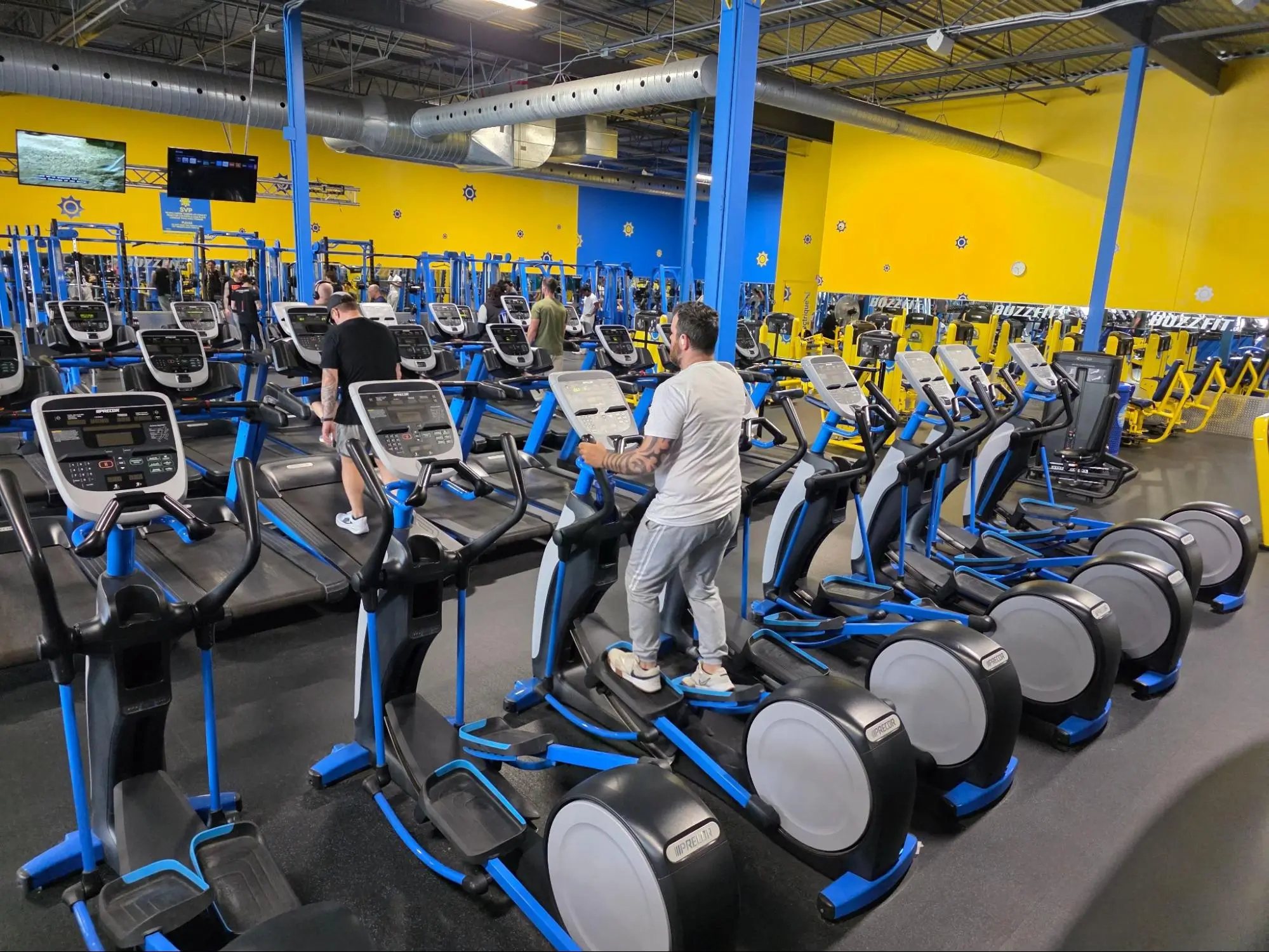 People exercising on elliptical machines and treadmills in a modern commercial gym.